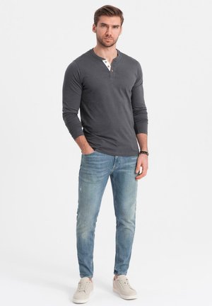 Mann trägt ein dunkelgraues Langarm-Henley-Shirt, hellblaue Jeans und beige Sneaker, steht mit einer Hand in der Tasche vor einem weißen Hintergrund.