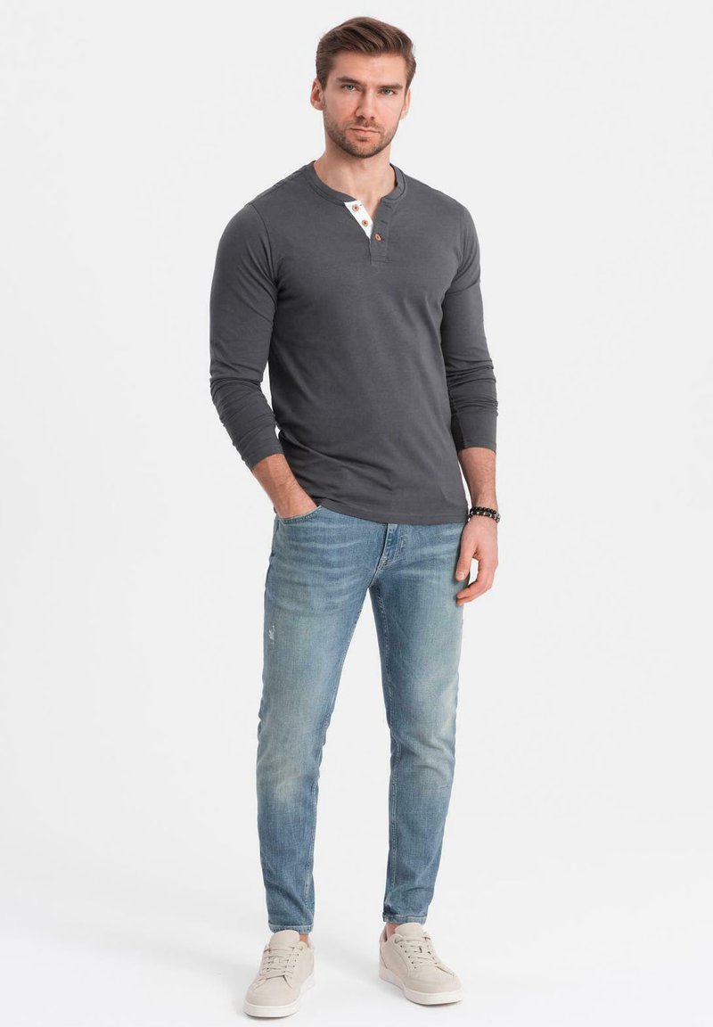 Man som bär en mörkgrå långärmad henley-tröja, ljusblå jeans, beige sneakers och står med en hand i fickan mot en vit bakgrund.