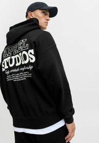 Schwarzer Hoodie mit großem weißen Textdesign auf dem Rücken, das "APPAREL STUDIOS" und zusätzlichen Text zeigt. Weicher Stoff, lockere Passform.