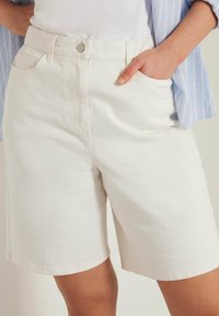 Valkoiset farkkushortsit, joissa on suora leikkaus, etutaskut ja napitus vyötäröllä. Sileä pinta, jossa on minimaalista ompelutietoa.