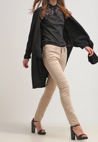 Svart blus med spetskrage, beigefärgade åtsittande byxor och en lång svart kappa. Modellen bär svarta klacksandaler. Slätt tyg, skräddarsydd passform.