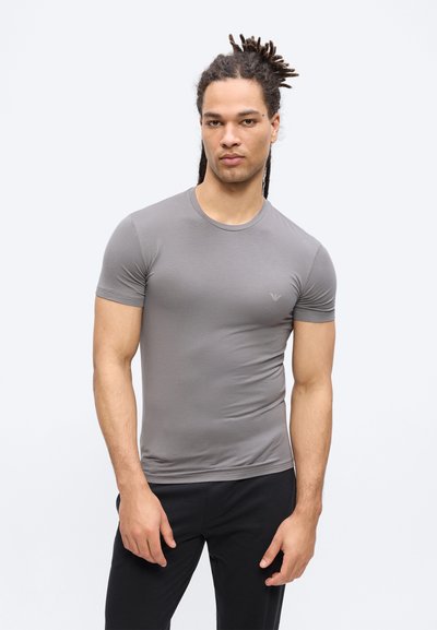 Άνδρας με dreadlocks που φοράει εφαρμοστό γκρι κοντομάνικο πουκάμισο και μαύρο παντελόνι, στέκεται μπροστά σε απλό ανοιχτό φόντο.