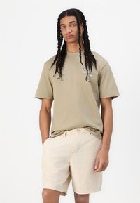 T-shirt in cotone khaki con maniche corte e vestibilità rilassata, abbinato a pantaloni corti in lino beige con chiusura a bottone e tasche laterali.