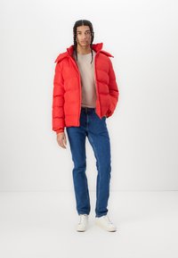Rød polstret jakke med høj krave, med lynlås lukning og raglanærmer, parret med blå jeans og hvide sneakers. Glat tekstur.