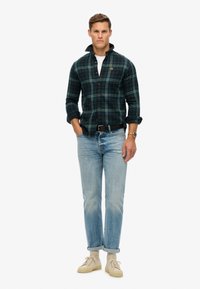 Groene en marineblauwe geruite flannel shirt met knoopsluiting, opgerolde mouwen, lichtblauwe rechte jeans en beige veterschoenen.