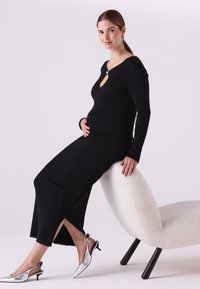 Čierne rebrované šaty s dlhým rukávom, výstrihom s otvorom a bočným rozparkom, doplnené striebornými sandálmi na zadnom popruhu. Modelka ležérne sedí na stoličke.