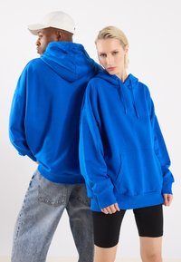 Blå overdimensjonerte hettegensere laget av sweatshirtstoff med kengurulommer, snørebånd og ribbestrikkede mansjetter, kombinert med denim og svarte shorts.