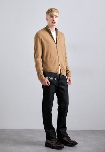 Jauns vīrietis ģērbj kamieļu krāsas jaku ar rāvējslēdzēju, melnās džinsās un brūnās kurpēs, turot melnu Emporio rokassomu pret vienkāršu pelēku fonu.