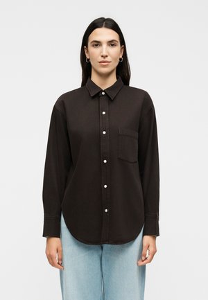 Kobieta o ciemnych, prostych włosach nosi czarną koszulę na guziki z lewą kieszenią na piersi oraz jasnoniebieskie dżinsy, stoi na jednolitym tle.