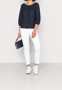 Blusa blu navy a trama con maniche a tre quarti, abbinata a jeans bianchi slim-fit e sneakers bianche. Tiene in mano una borsa nera con accenti rossi e bianchi.