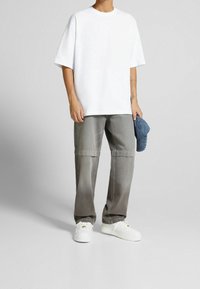 Person som bär en överdimensionerad vit t-shirt, lösa grå byxor, vita sneakers och håller en blå denimkeps i höger hand.