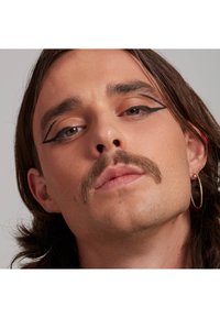 Nahaufnahme eines Gesichts mit langen braunen Haaren, das sich durch einen kräftigen schwarzen Eyeliner, einen ausgeprägten Schnurrbart und einen Creolenohrring auszeichnet, vor einem hellen Hintergrund.