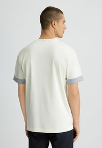 Camiseta de algodón en color crema, de manga corta, con puños a rayas grises y cuello redondo. Corte recto con una textura suave. Sin impresiones visibles.