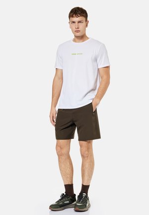 Junger Mann steht, trägt weißes "KEEP ACTIVE" T-Shirt, dunkle Shorts, braune Socken und grüne Sportschuhe vor einfarbigem Hintergrund.