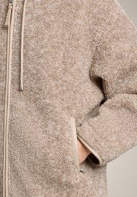 Beżowa pluszowa kurtka z zamkiem błyskawicznym i sznurkiem. Posiada przyjemną w dotyku fakturę, boczne kieszenie z beżowym wykończeniem oraz metalowe akcenty.