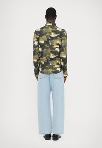 Camouflage-patterned jacket in green, beige, and black, with a high collar, long sleeves, and contrasting detailing. Paired with light blue wide-leg jeans.
