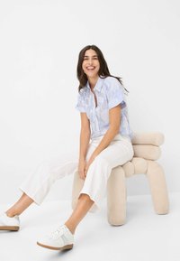 Hellblaues Button-Up-Hemd mit floralem Muster, kombiniert mit weißen 3/4-Hosen und weißen Sneakern, sitzt auf einem beigen runden Hocker.