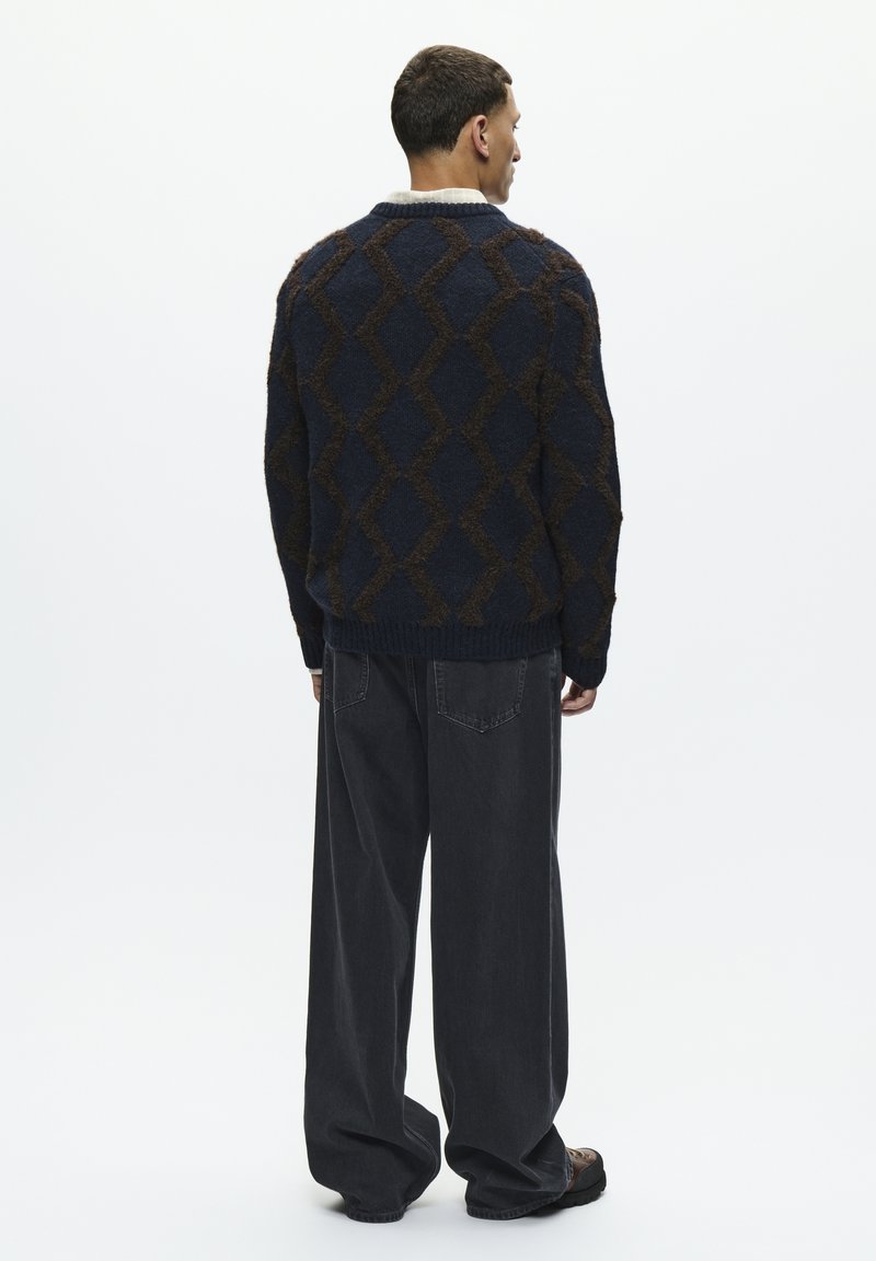 Pull marine avec un motif en losanges texturé marron, associé à un pantalon noir ample. La tenue présente une coupe décontractée et des poignets côtelés.