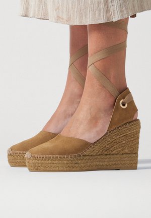 Espadrillas a zeppa in camoscio tan con suole in corda intrecciata e cinturini che si avvolgono intorno alla caviglia, indossate con una gonna beige.
