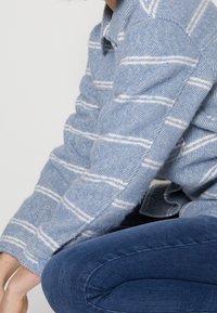 Person som bär en blå och vit randig ulljacka och mörkblå jeans, sittande med en arm vilande på sitt knä.