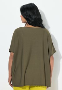Olivově zelený, volně střižený top s širokým výstřihem a krátkými rukávy. Tkanina je strukturovaná, v kombinaci se žlutými kalhotami pro kontrast.