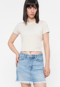 Kobieta z prostymi ciemnymi włosami, ubrana w kremowy krótki T-shirt i jasnoniebieską dżinsową spódnicę z postrzępionym brzegiem, stojąca na tle białym.