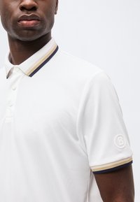 Κοντινό ενός άνδρα που φοράει λευκό πόλο πουκάμισο με γιακά και μανσέτες μπεζ και ναυτικού μπλε ριγέ, με στρογγυλό λογότυπο στο μανίκι.