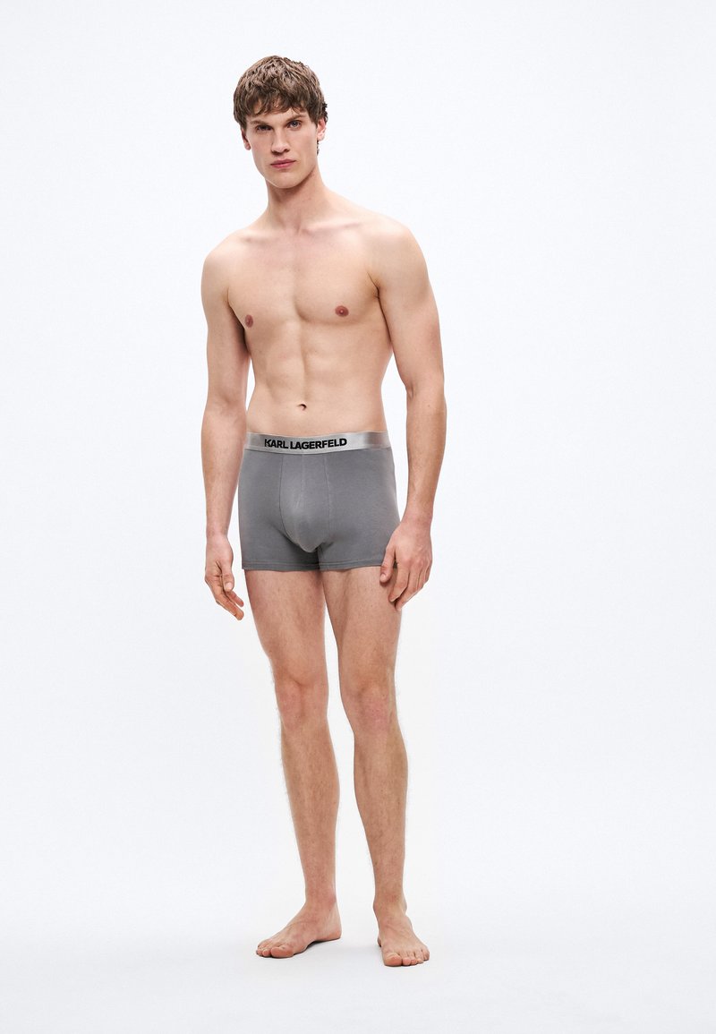 Graue Boxershorts aus dehnbarem Material mit einem schwarzen Bund, der das Logo "KARL LAGERFELD" trägt. Das Modell steht barfuß.
