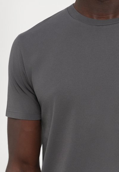 Close-up van een persoon die een effen grijze T-shirt met korte mouwen en ronde hals draagt met een gladde stofstructuur.