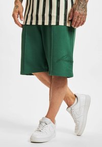 Shorts en coton verts avec un logo brodé, associés à des baskets en cuir blanches. Ils présentent une coupe décontractée et une texture lisse.