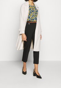 Cappotto trench beige sopra un top floreale blu, pantaloni neri cropped e décolleté neri con design a fantasia.