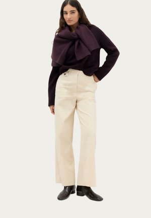 Kvinde iført mørkelilla sweater med matchende sweater draperet over skuldrene, beige vidde bukser og sorte ankelstøvler, der står med hånden i lommen.