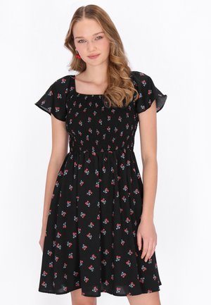 Nainen, jolla on pitkät laineikkaat hiukset ja musta mekko, jossa on punavalkoinen kukkakuosi ja liehukehihat, seisoo valkoista taustaa vasten.