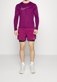 Lila langärmliges Sportoberteil mit einem Nike-Logo, kombiniert mit passenden lila Shorts. Trägt dazu graue Sportschuhe und weiße Socken.