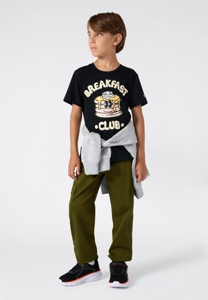 Jongen met bruin haar staat naar links te kijken, draagt een zwarte "Breakfast Club" T-shirt, olijfkleurige cargobroek, zwarte sneakers en een grijze sweater om zijn taille geknoopt.