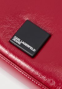 Melnas kvadrātveida Karl Lagerfeld Jeans logotips uz sarkanas ādas ar spīdīgu tekstūru, tuvplāns.