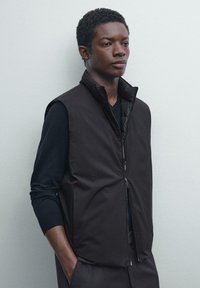 Jeune homme portant un gilet rembourré noir zippé sur une chemise à manches longues noire, se tenant avec une main dans sa poche contre un mur clair et uni.