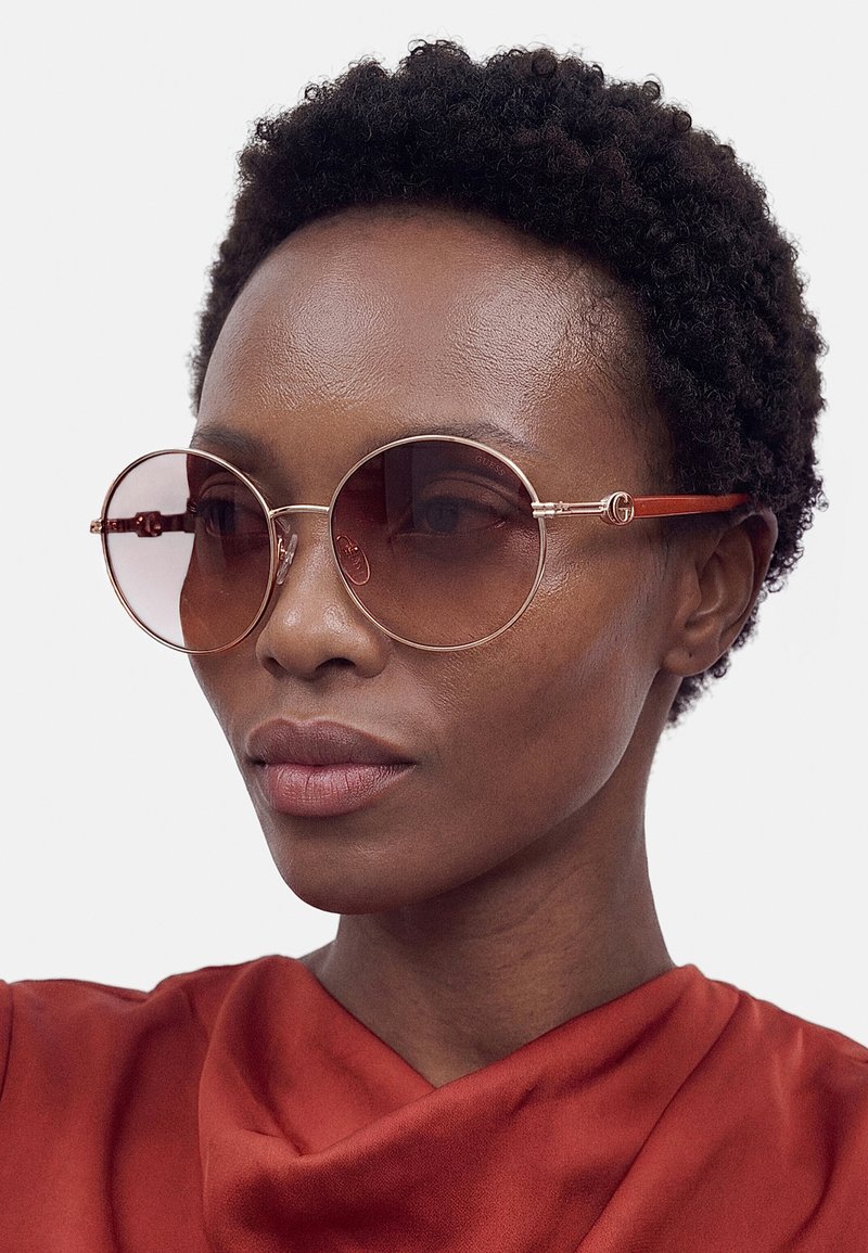 Femme aux cheveux courts et bouclés portant de grandes lunettes de soleil rondes avec des montures fines en or rose et un haut rouge sur un fond clair uni.