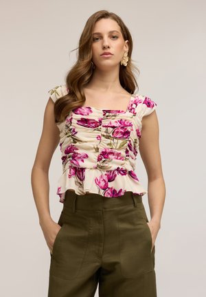 Jeune femme aux longs cheveux ondulés porte un haut floral froncé avec des fleurs roses et un pantalon vert olive, debout les mains dans les poches.