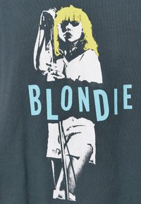 Černé tričko s grafickým potiskem osoby se žlutými vlasy a mikrofonem. Text říká "BLONDIE" v světle modré barvě.