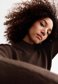 Maglione marrone a coste con scollo alto, indossato da una persona con capelli ricci. La texture sembra morbida e accogliente.