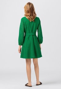 Grön spetsklänning med texturerad mönster, belagd midja och långa puffärmar som slutar ovanför knäet. Platta svarta sandaler fulländar looken.