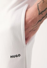 Hvide bomulds shorts med en sidelomme, der har et sort "HUGO" logo på benet. Stoffet ser glat ud med en let glans.