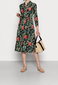 Vestito midi floreale in tessuto verde scuro con rose rosse e motivi di foglie, vita aderente, maniche lunghe e colletto. Tiene una borsa di paglia.