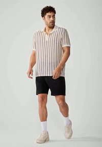 Kortärmad pikétröja med vertikala ränder i beige, marinblå och krämfärgad; mjukt tyg; kombinerad med svarta shorts och ljusa sneakers.