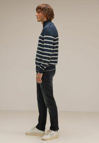 Marineblauwe trui met lichtgrijze horizontale strepen, ritssluiting bij de kraag, gecombineerd met donkere jeans en witte sneakers. Eenvoudig, sportief design.
