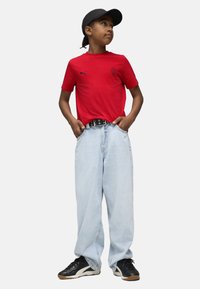 Röd bomulls T-shirt med korta ärmar och en logotyp, ihop med ljusblå baggy jeans och svarta sportskor. Svart keps bärs bakåt.