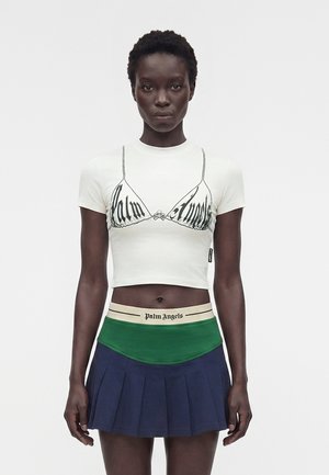 Γυναίκα με κοντά μαλλιά που φοράει λευκό crop top με μαύρο τύπωμα σουτιέν και κείμενο, πράσινη και μπεζ ζώνη μέσης, και μπλε πλισέ μίνι φούστα, που κοιτάει μπροστά.