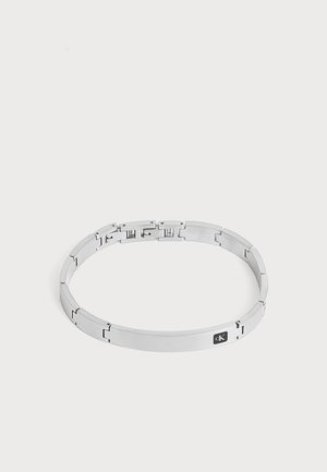 Bracelet en métal argenté avec plaque rectangulaire et petite étiquette noire avec logo sur fond clair uni.
