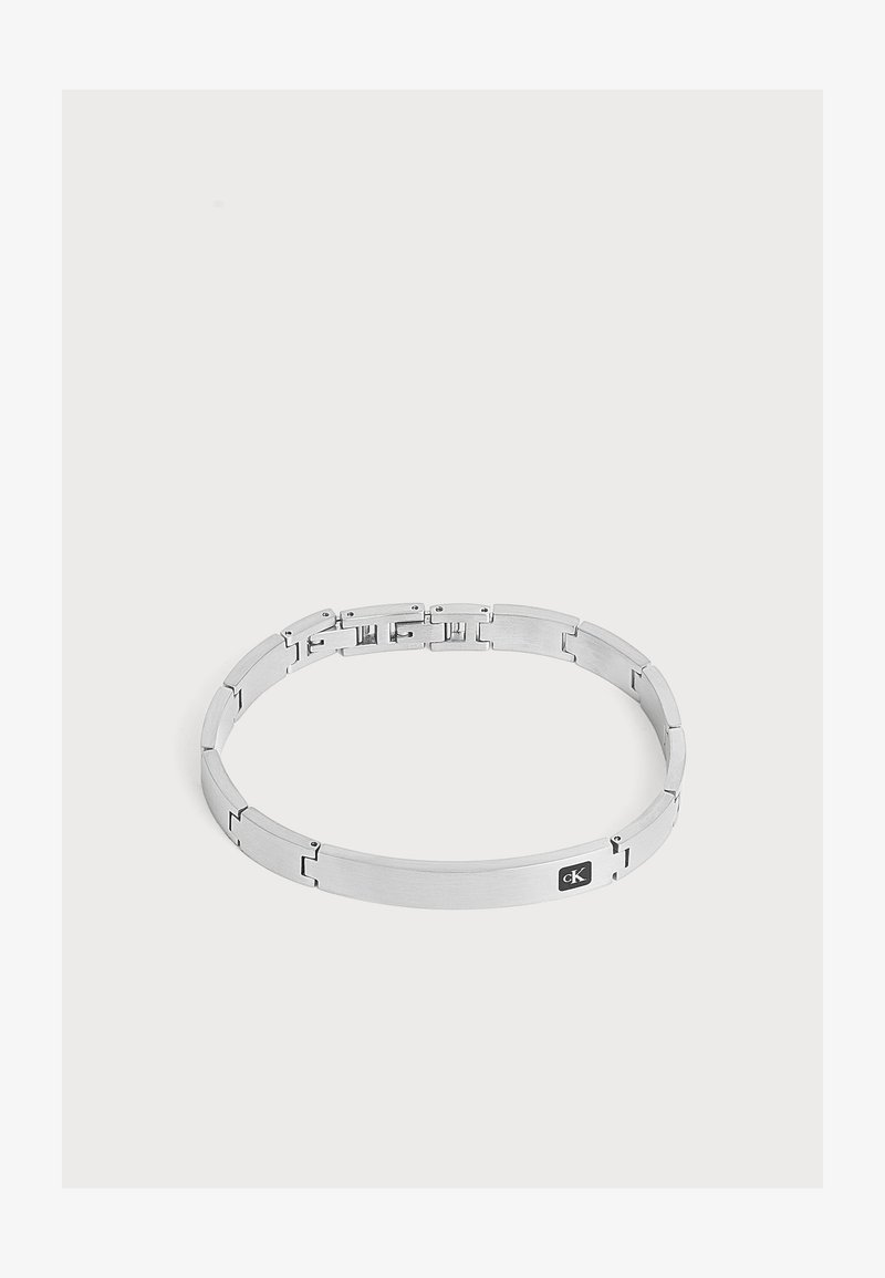 Bracelet en métal argenté avec plaque rectangulaire et petite étiquette noire avec logo sur fond clair uni.